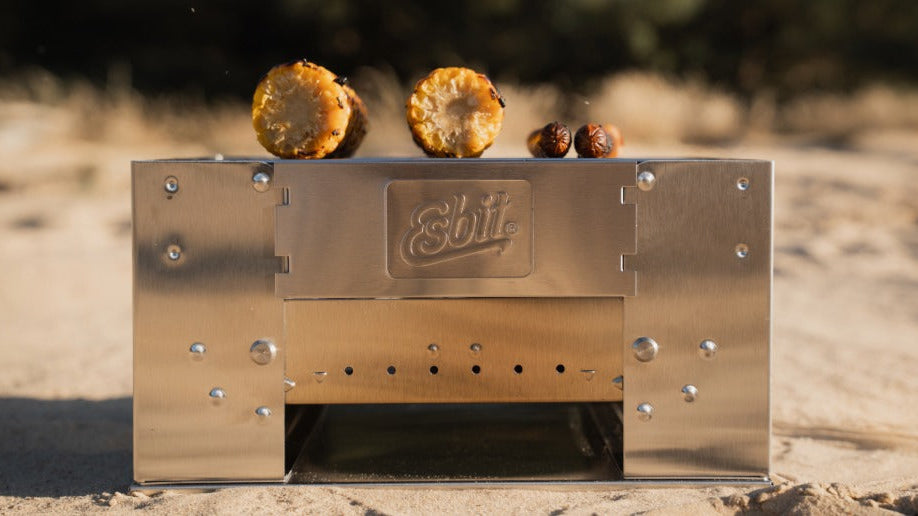 Kompakter Esbit Edelstahl-Holzkohlegrill BBQ300S im Einsatz am Strand, mit zwei Maiskolben und zwei Würstchen auf dem Rost.