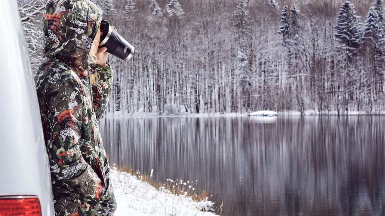 Junge Frau in Winterjacke steht angelehnt an einem Auto an einem verschneiten See im Wald und trinkt aus einem Esbit Thermobecher.