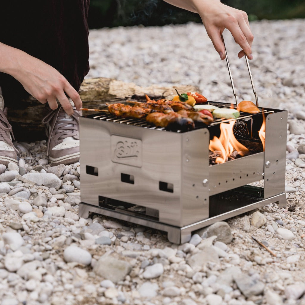 Person grillt Gemüse und Grillspieße auf einem Esbit Edelstahlgrill mit offenem Feuer in einer Outdoor-Umgebung.