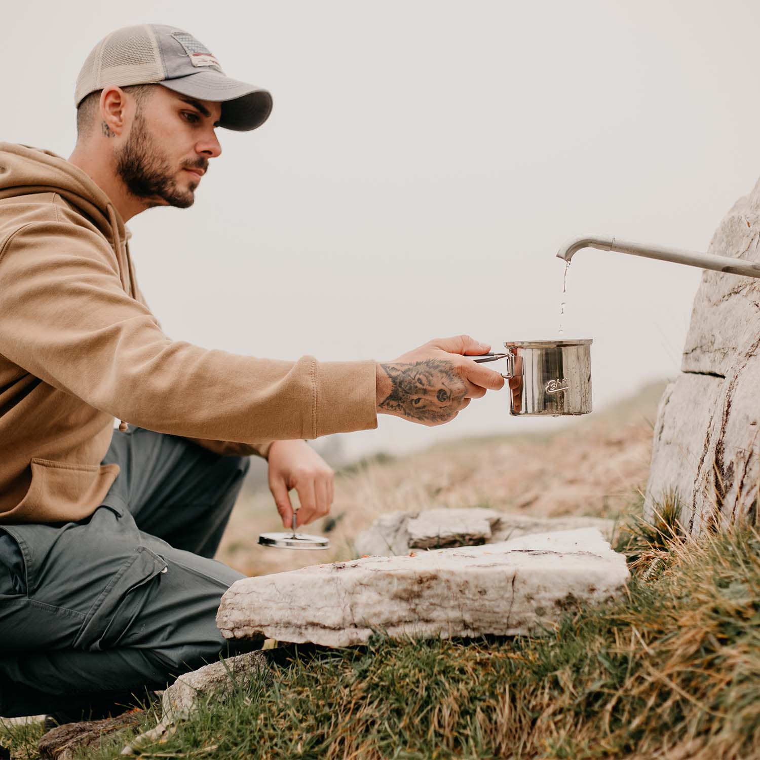 Mann mit beiger Mütze füllt Edelstahl Camping Topf mit Wasser aus einem Brunnen.