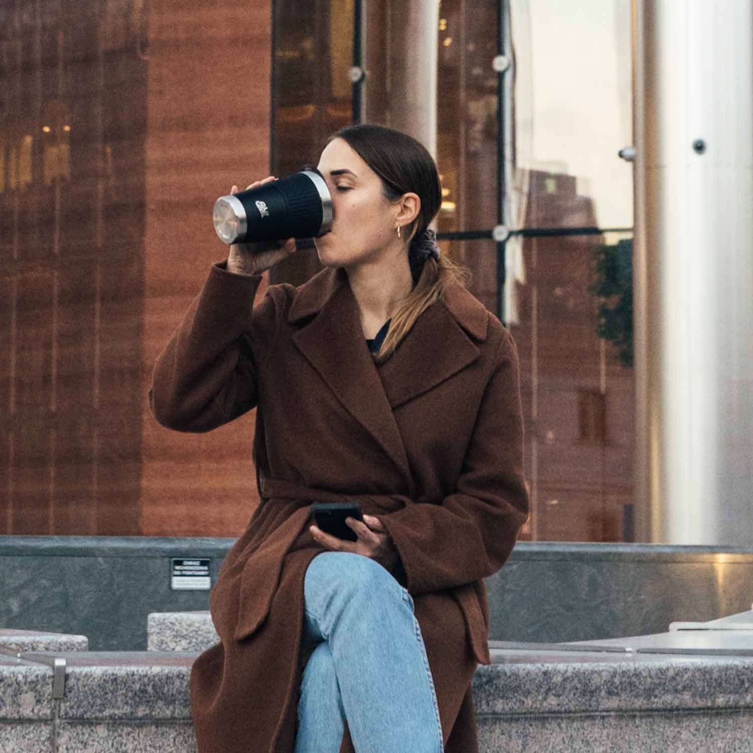 Frau sitzt in der Stadt an einem Brunnen und trinkt aus ihrem SCULPTOR Thermobecher in Schwarz.