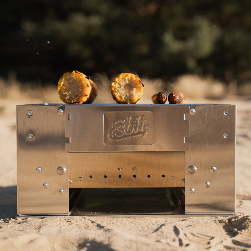 Esbit Edelstahlgrill BBQ300S mit gegrilltem Maiskolben und Würstchen in einer Outdoor-Umgebung.