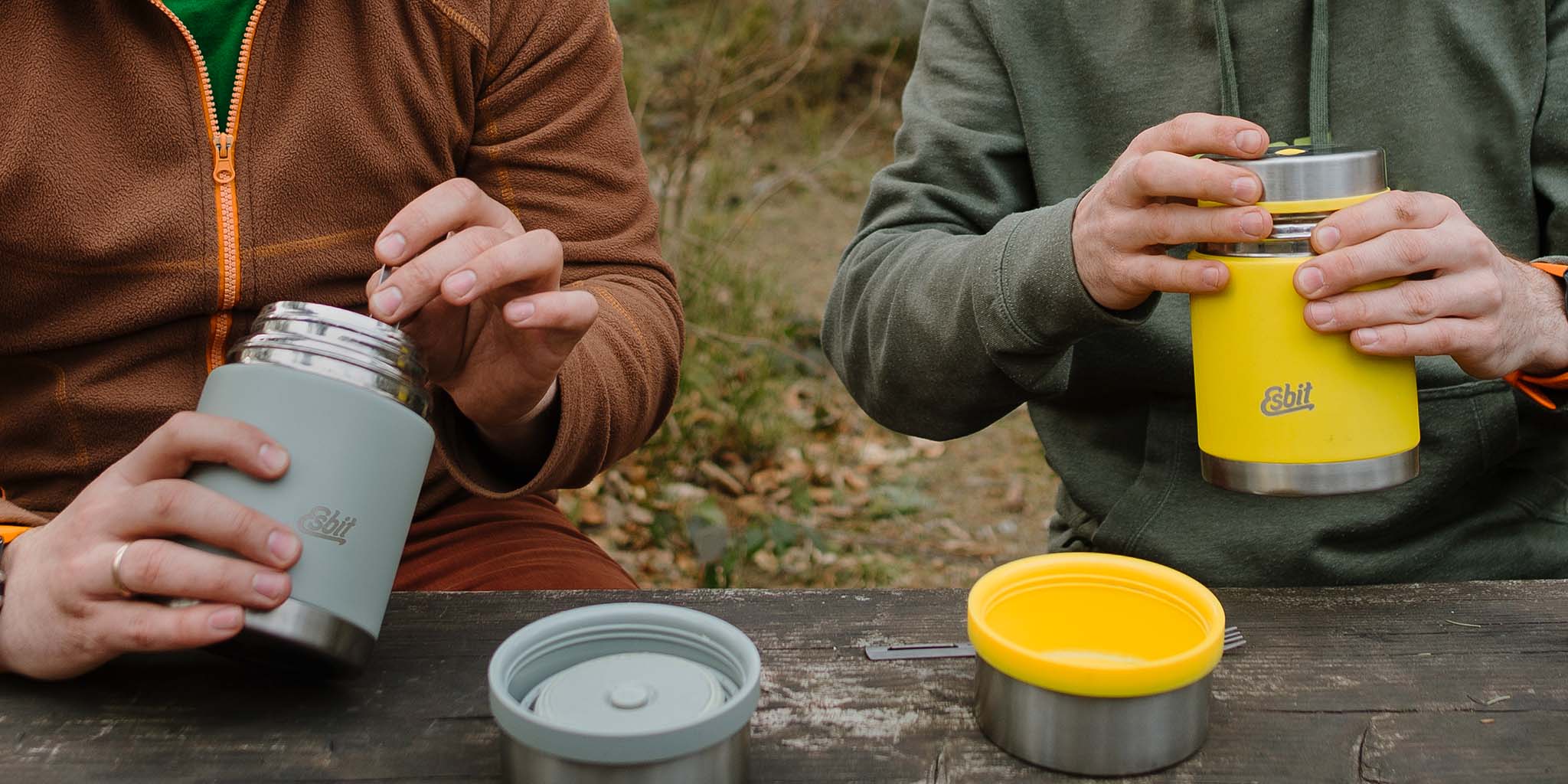 Zwei Männer essen unterwegs an einem Holztisch aus ihren SCULPTOR Thermobehältern in Grau und Gelb.