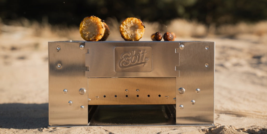 Kompakter Esbit Edelstahl-Holzkohlegrill BBQ300S im Einsatz am Strand, mit zwei Maiskolben und zwei Würstchen auf dem Rost.