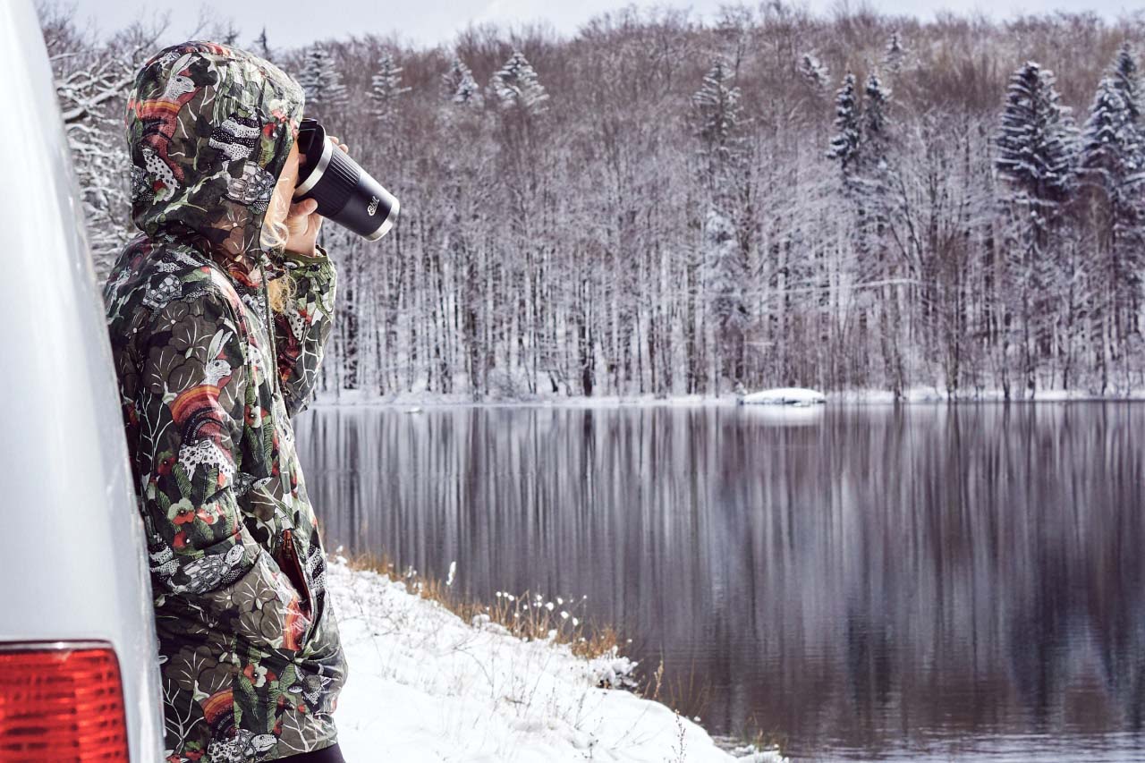 Junge Frau in Winterjacke steht angelehnt an einem Auto an einem verschneiten See im Wald und trinkt aus einem Esbit Thermobecher.