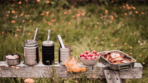 Drei geoeffnete Esbit Thermobehaelter, eine Schale Erdb auf einer Holzbank in der Natur.