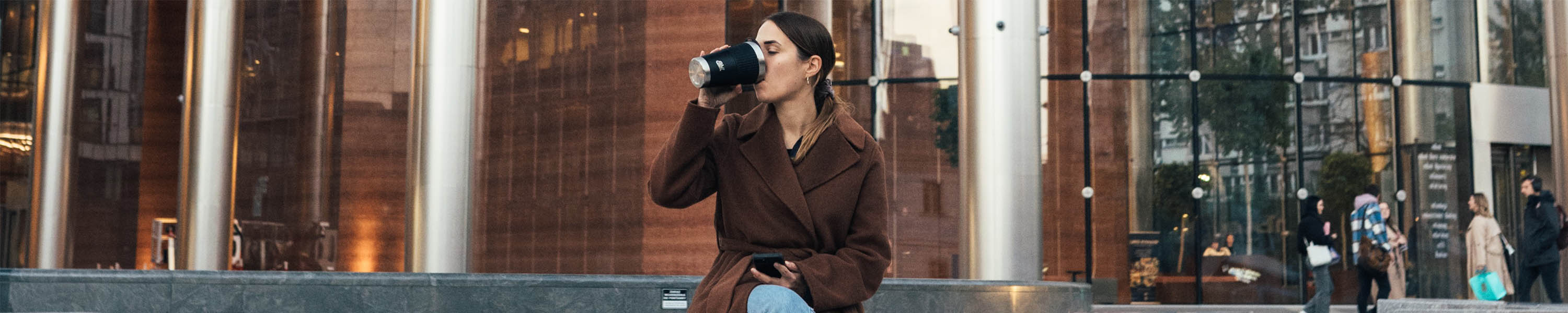 Frau sitzt vor einem Gebäude und trinkt aus dem SCULPTOR Thermobecher mit Manschette in Schwarz