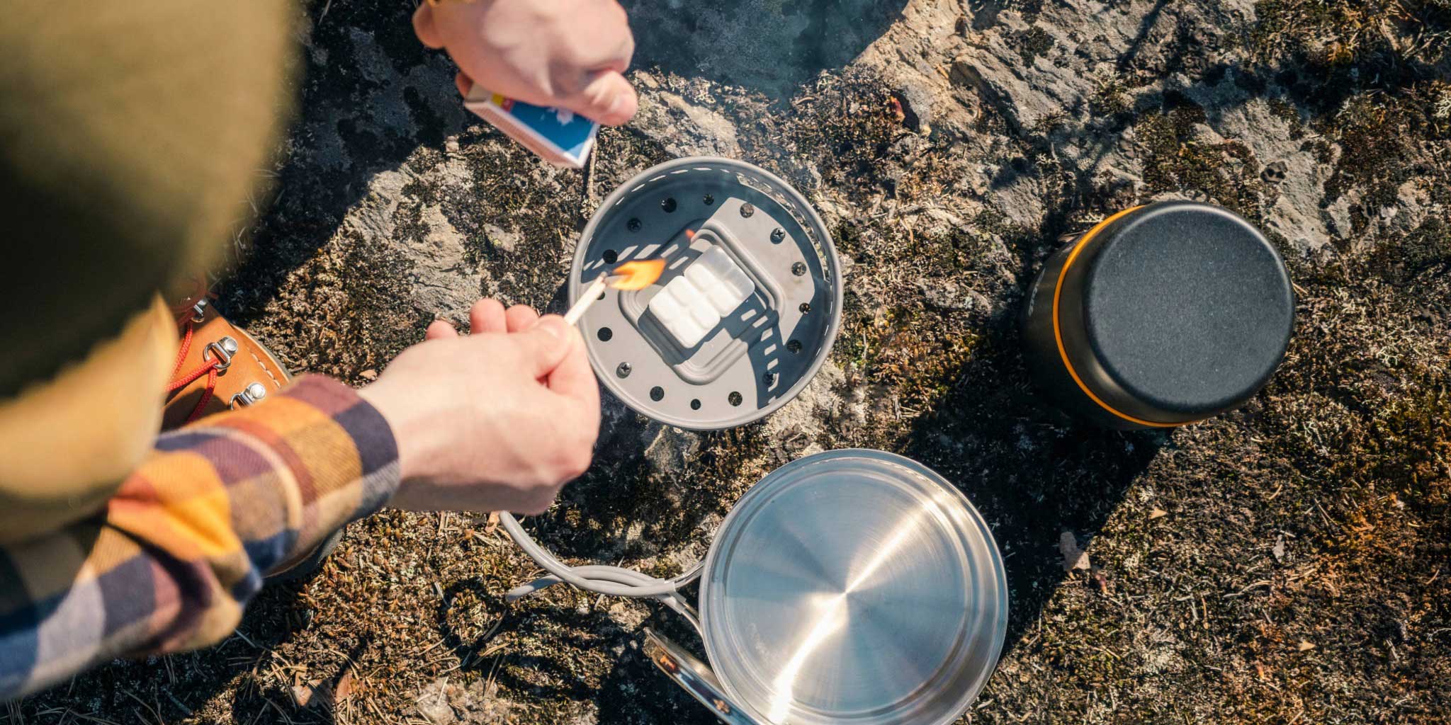 Esbit Outdoor-Kocher mit Trockenbrennstoff-Tabletten wird auf einem Felsen mit einem Streichholz entzündet. Daneben stehen ein Edelstahltopf und Food Jug