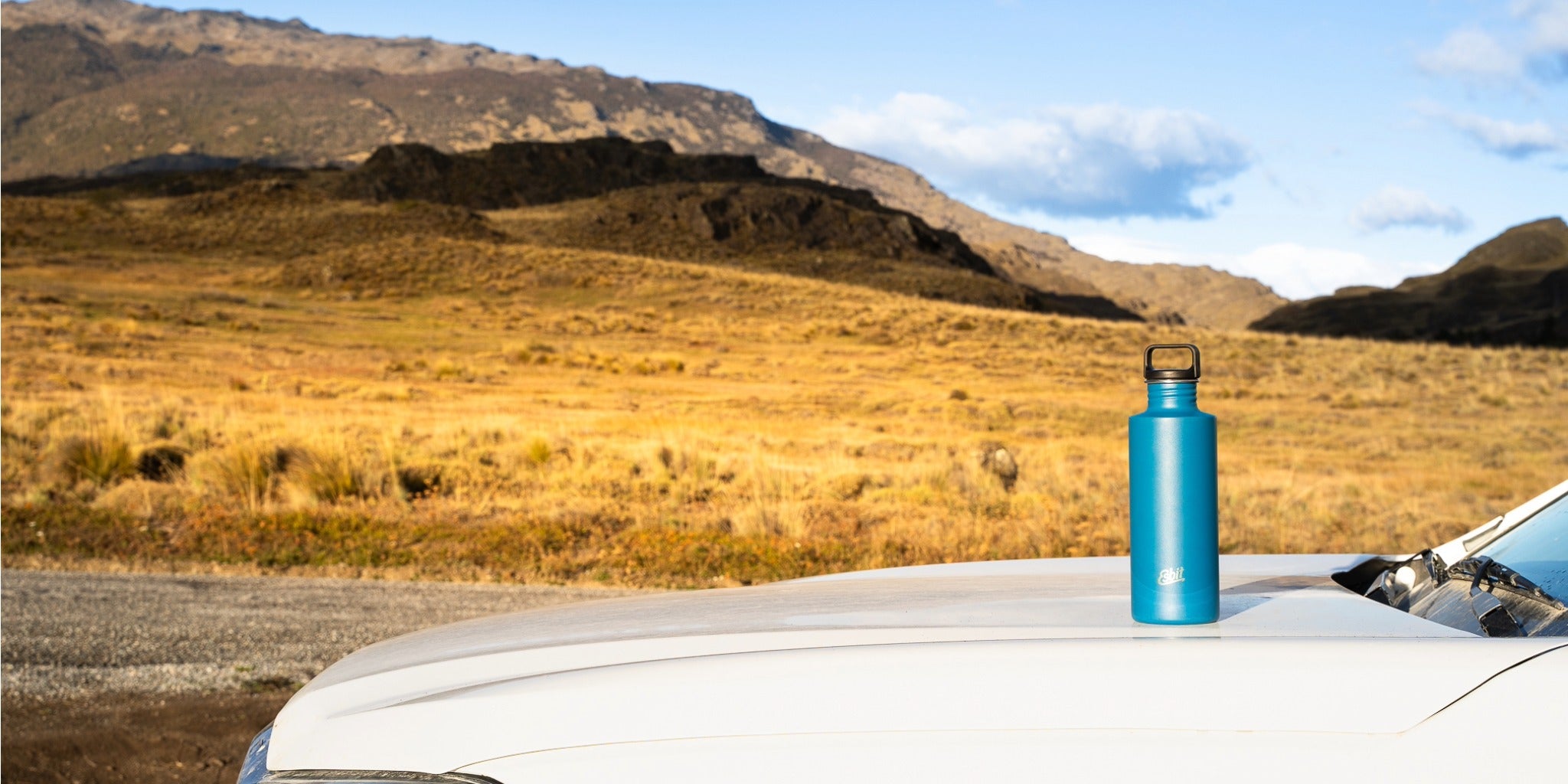 Esbit Edelstahl-Trinkflasche in Blau auf einem Fahrzeugdach vor einer weiten Berglandschaft.