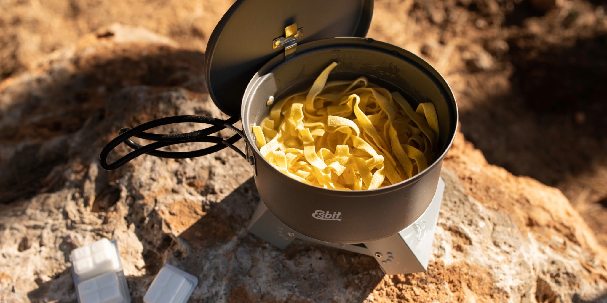 An Esbit cooking set with an open saucepan and pasta on an Esbit pocket stove in an outdoor setting.